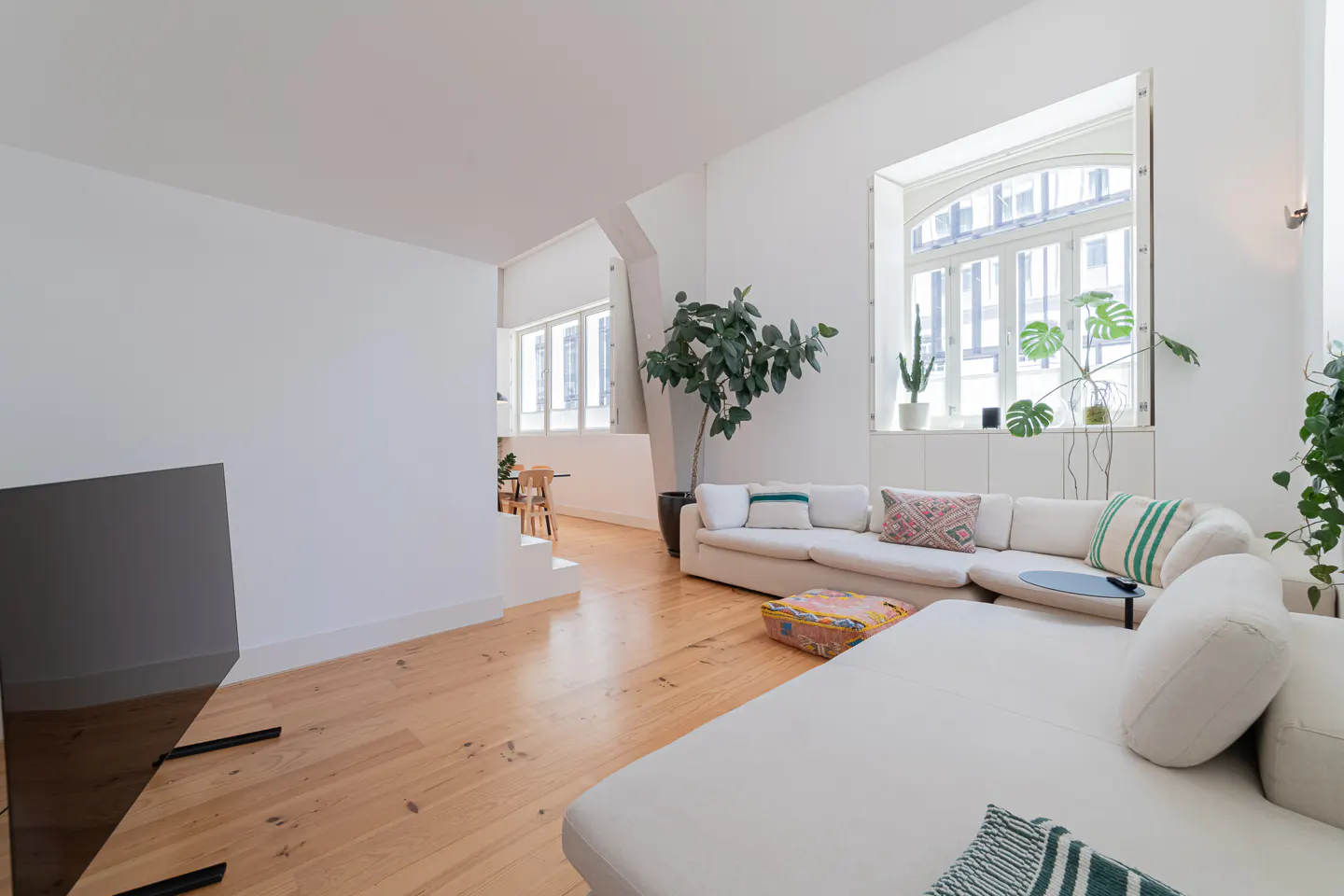 Bright, modern living room with white walls, wood floors, and a large white sectional sofa. Plants add greenery, and natural light streams through a large window.