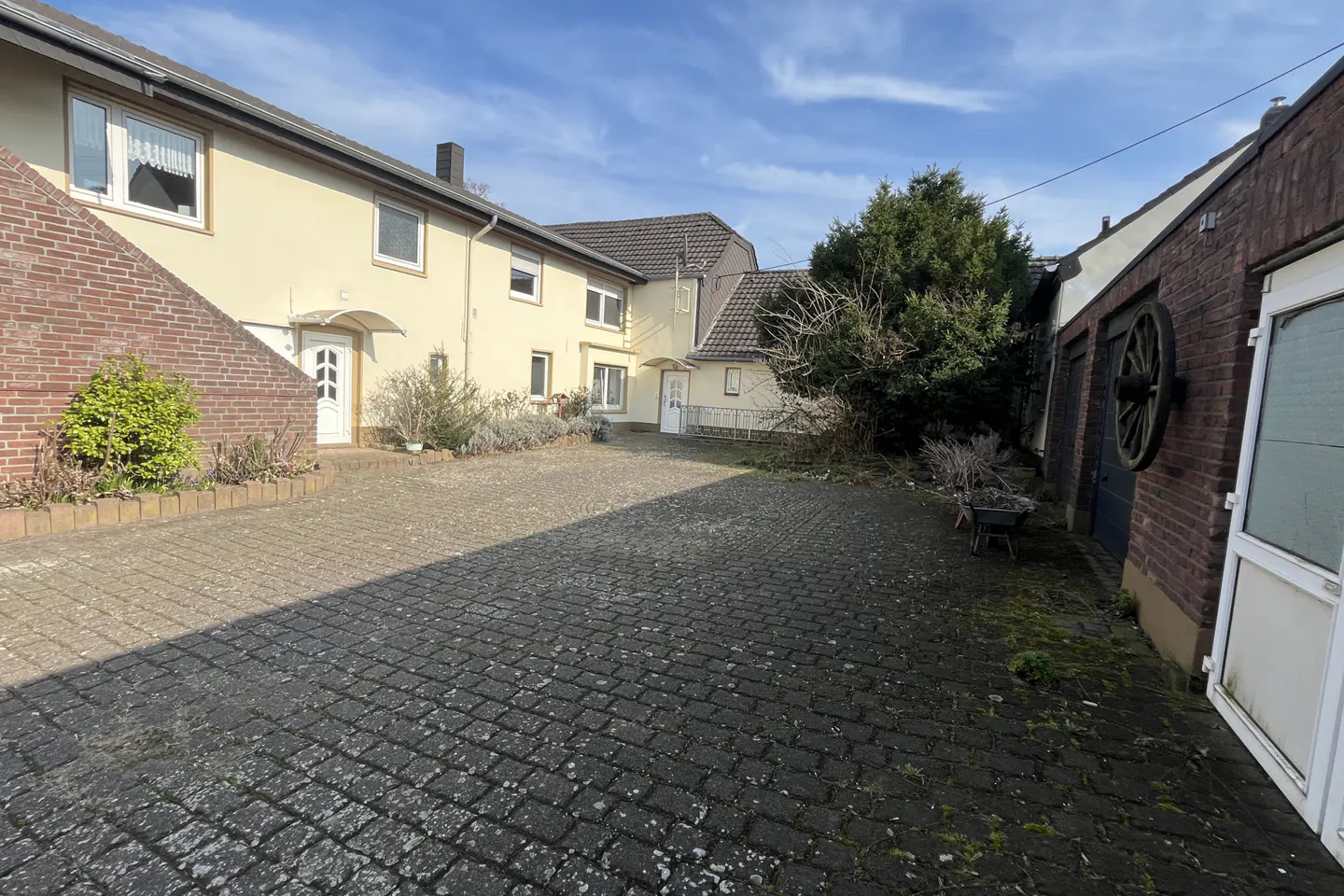Exterior view of a yellow two-story house with white doors and windows, a brick wall, and a cobblestone driveway.