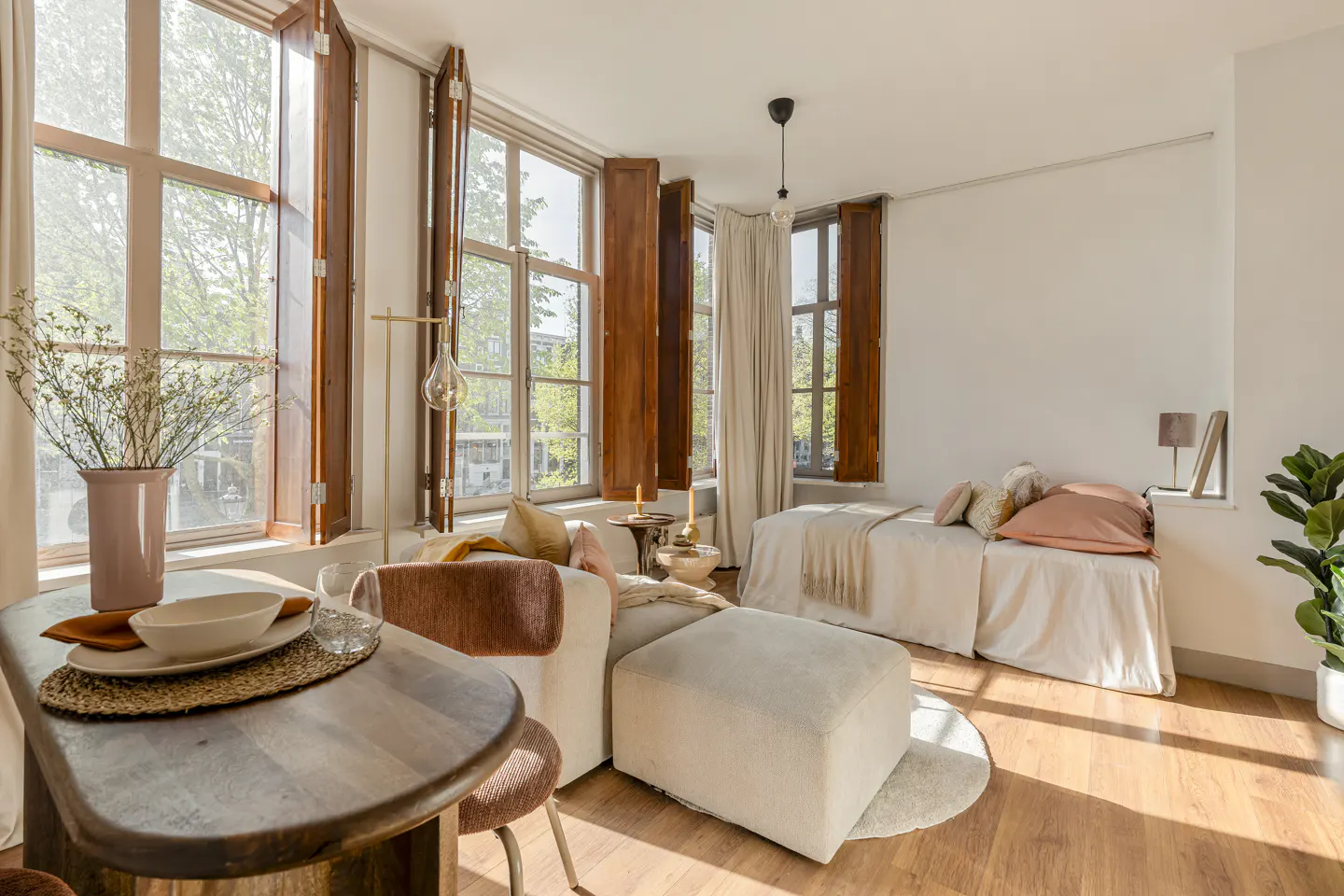Bright studio apartment with hardwood floors, large windows with wooden shutters, a bed, a sofa, and a dining table.