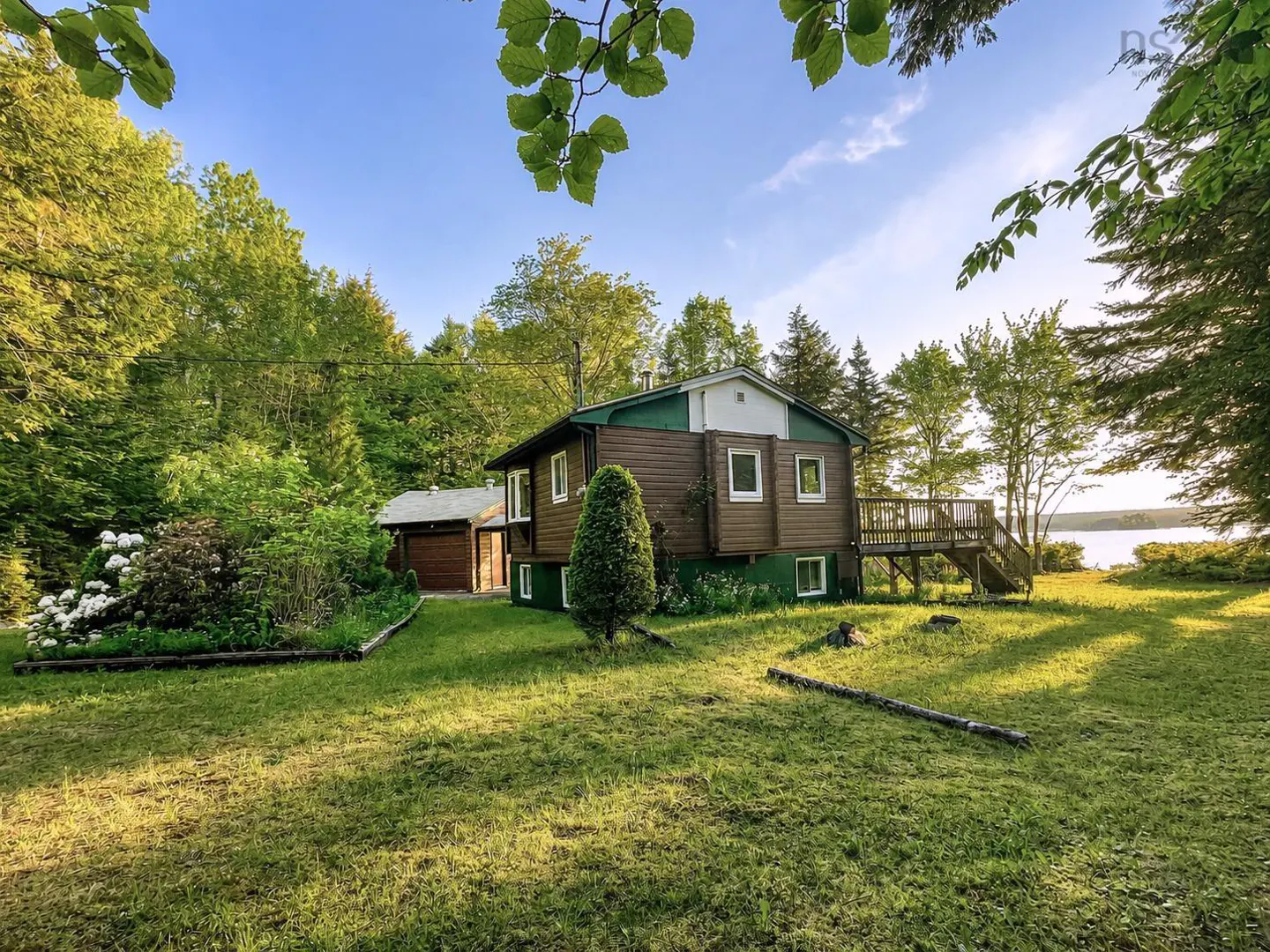 Brown house with green trim and a wooden deck, situated on a grassy lot surrounded by trees near a lake at sunset.