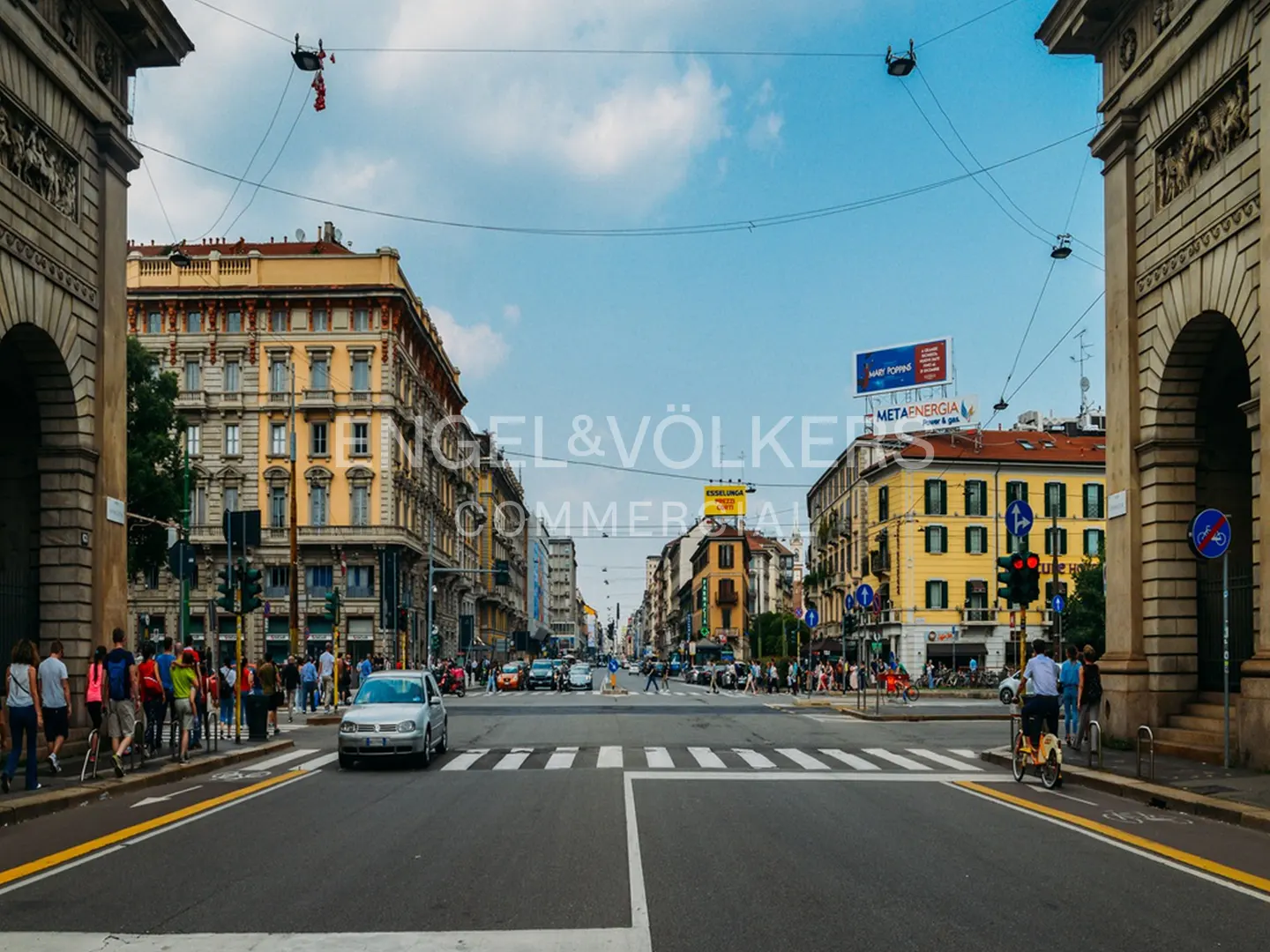 City street view with cars, pedestrians, and yellow buildings under a blue sky. Arches frame the scene.