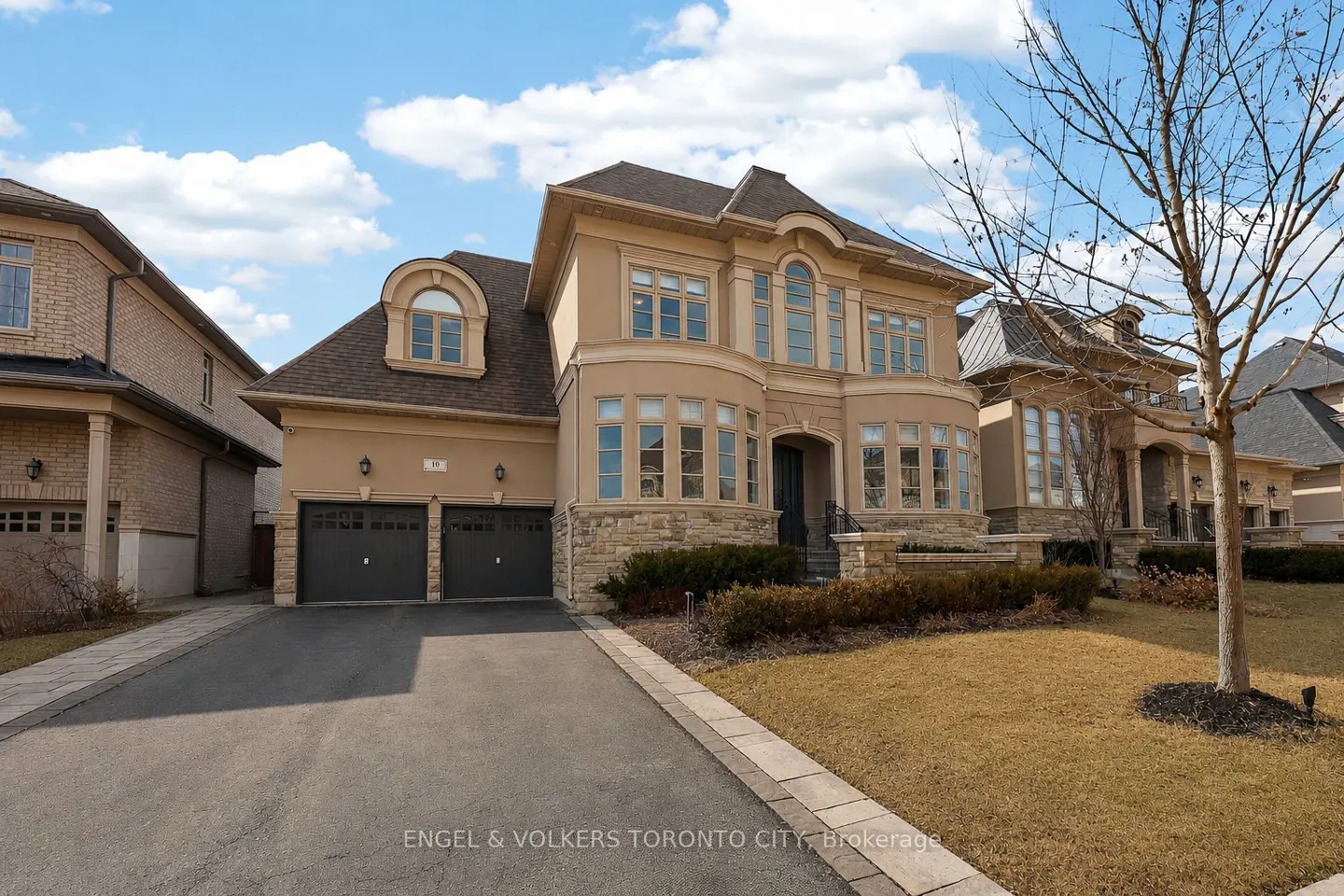 Two-story beige house with a dark roof, two-car garage, and a long driveway on a sunny day.