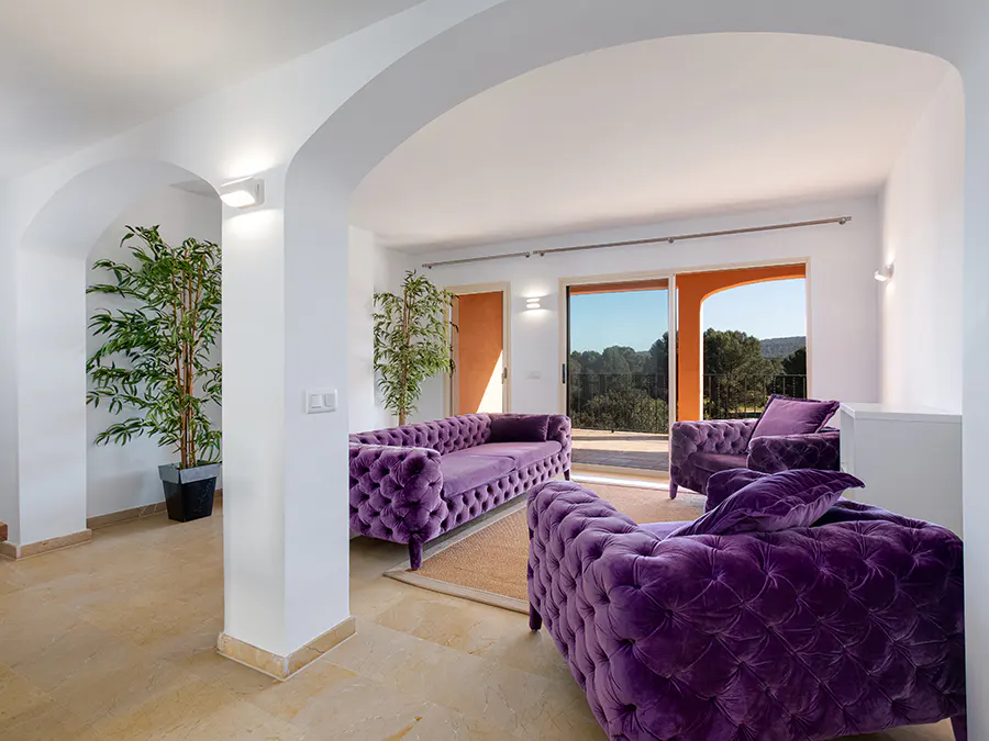 Bright living room with arched doorways, purple velvet sofas, and a view of trees through sliding glass doors.