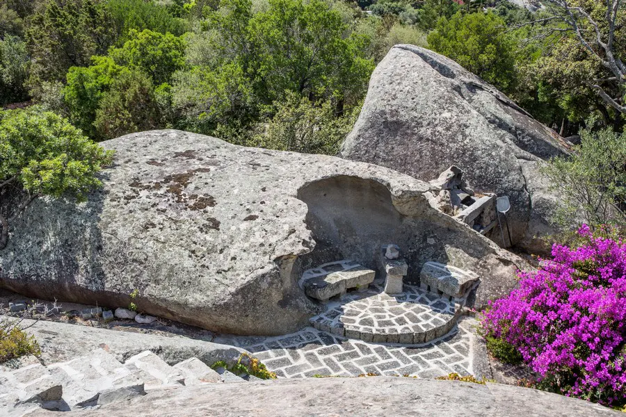 Outdoor seating area carved into a large rock formation, with stone benches, a brick patio, and purple flowers.