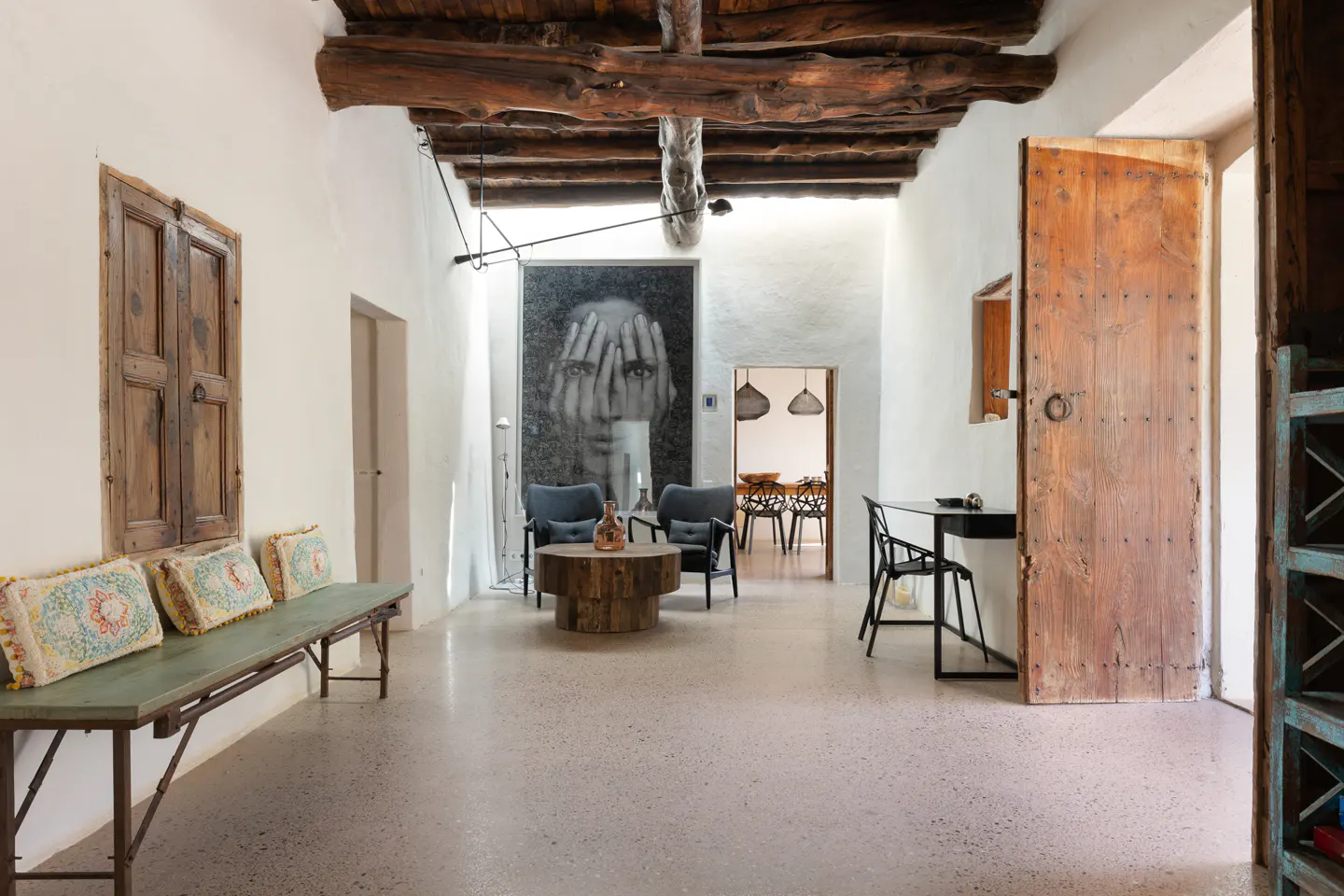 Bright, airy living room with exposed wood beams, white walls, and a large portrait. A wooden bench with pillows sits near a closed wooden window.