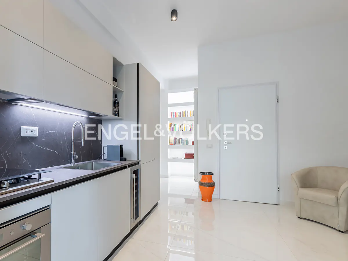Modern kitchen with light grey cabinets, dark marble backsplash, and a white door leading to a hallway with a bookshelf.