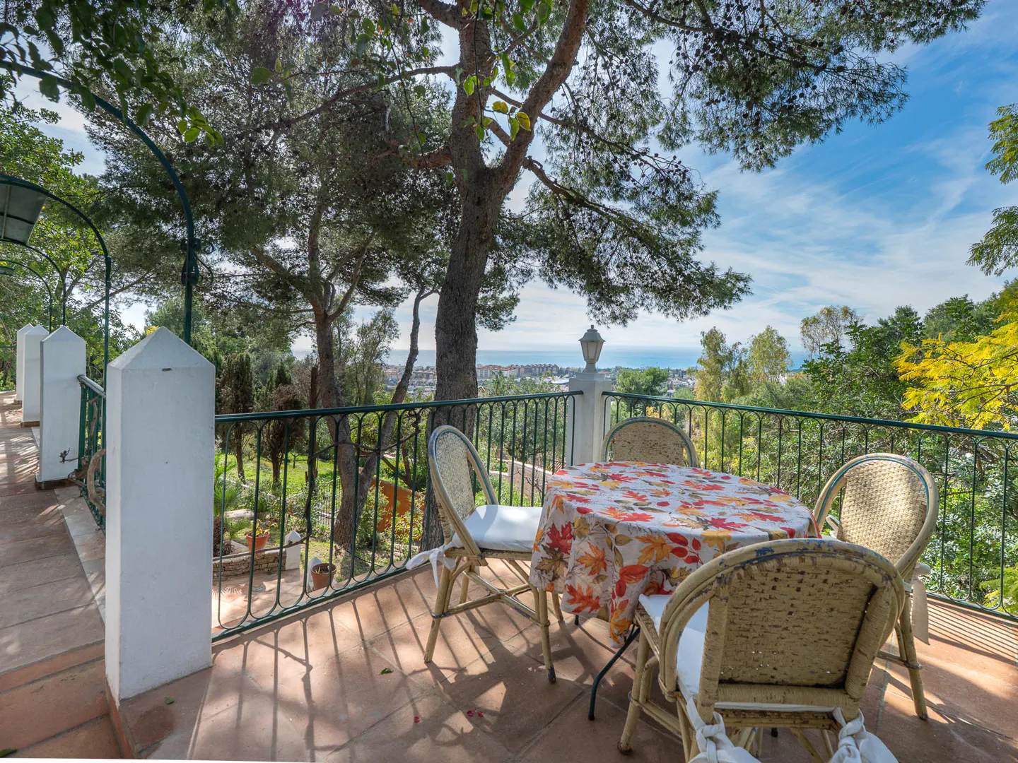 Outdoor patio with a round table covered with a floral tablecloth and four chairs overlooking a garden and ocean view.
