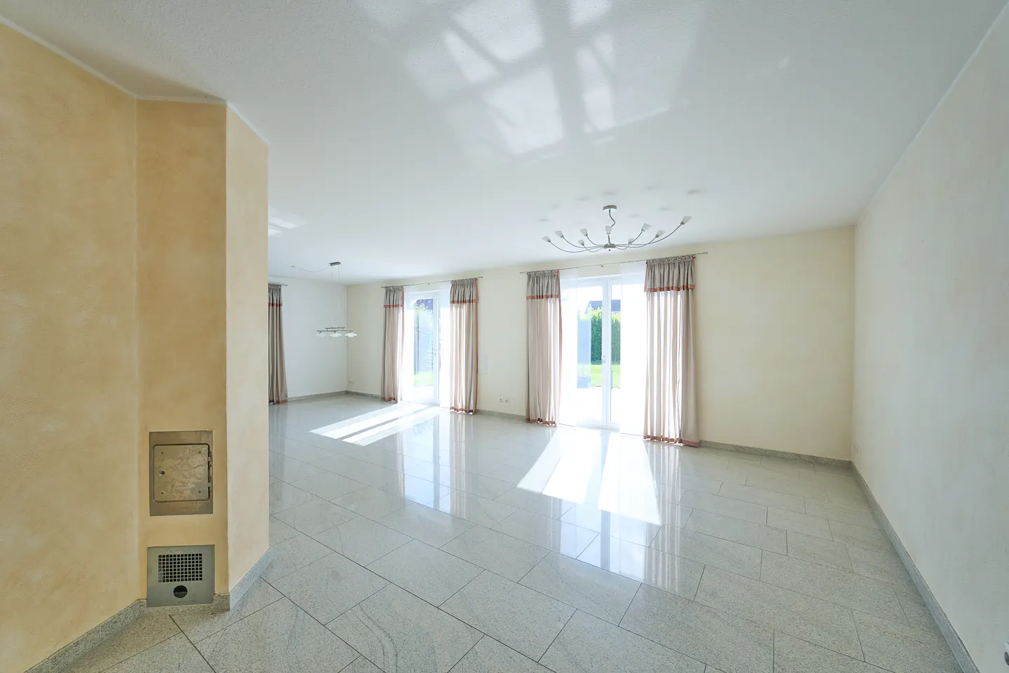 Spacious, bright living room with light-colored tile floors, beige walls, and large glass doors with curtains.