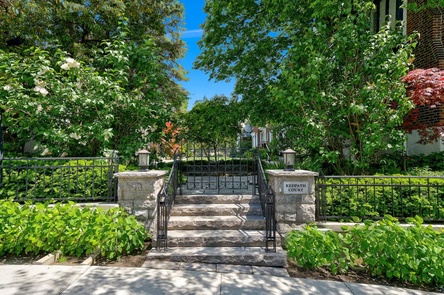 Stone steps lead to a black iron gate marked "Redpath Court," framed by lush greenery under a bright blue sky.