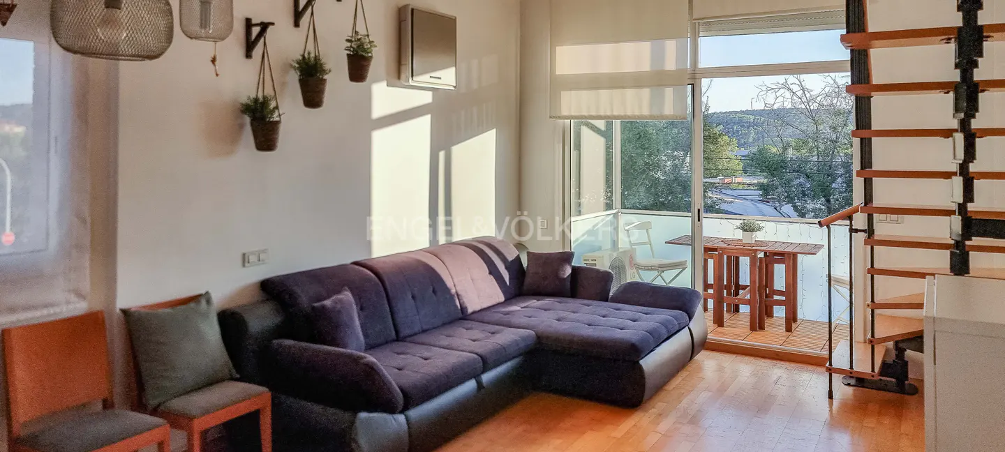 Living room with a dark blue sectional sofa, wooden chairs, and a balcony with a table and chairs. A spiral staircase is on the right.