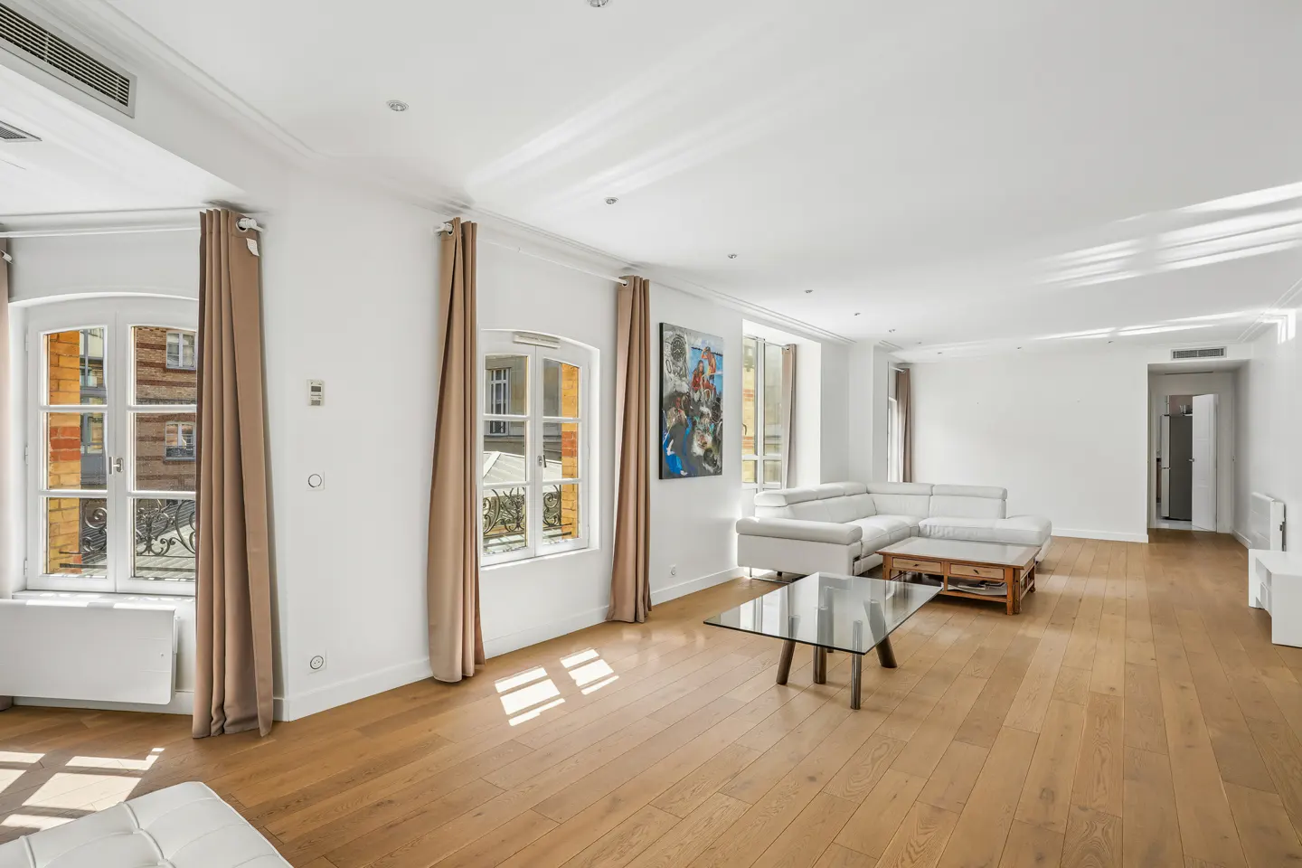 Bright, airy living room with hardwood floors, white walls, and large windows with tan curtains. A white sectional sofa and glass coffee table are centered.