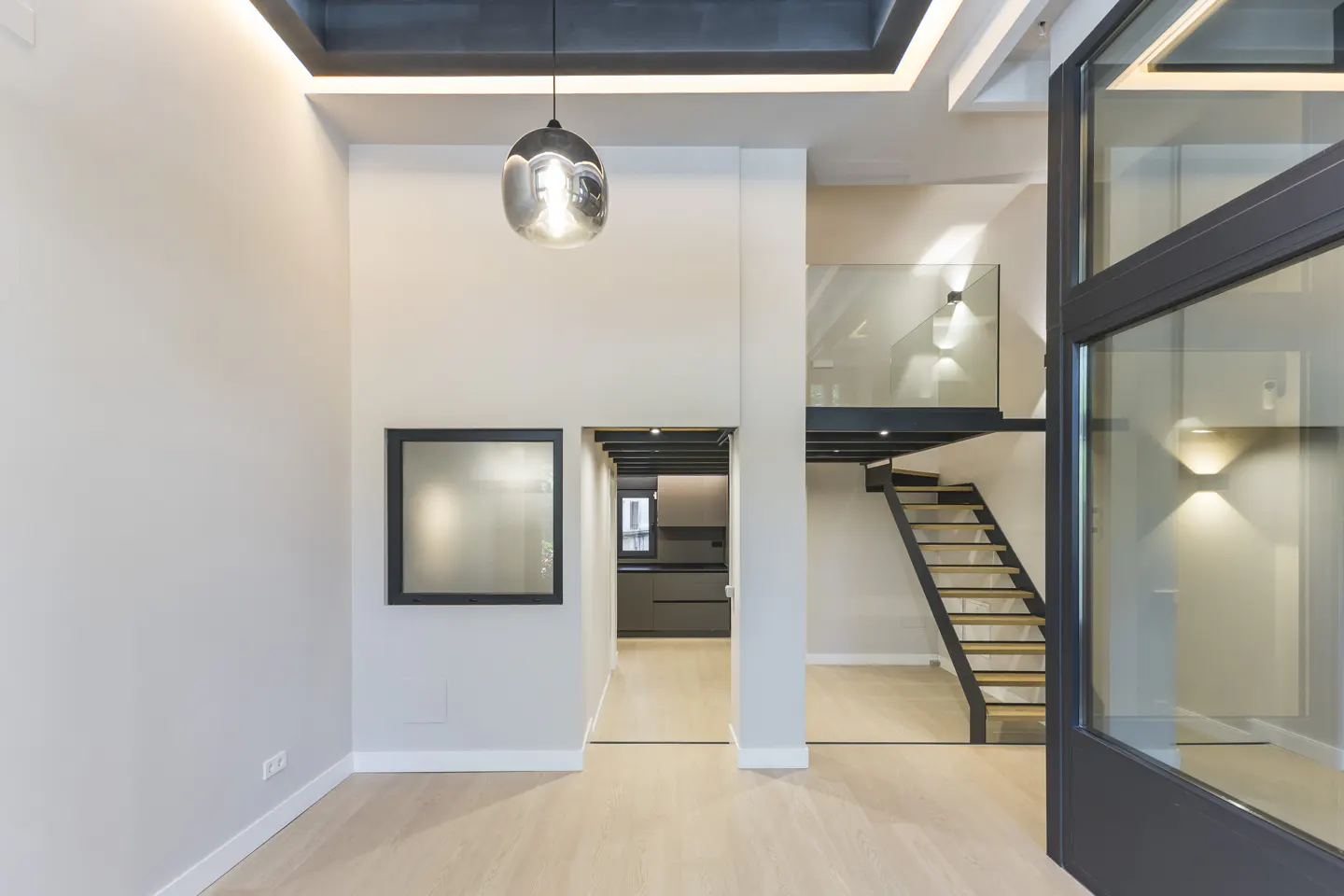 Modern interior with light wood floors, a glass-railed staircase, a pendant light, and a view into a dark kitchen.