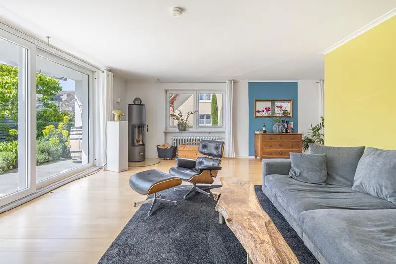Bright living room with a gray sofa, wood coffee table, black chair, and fireplace on a light wood floor with a large window.