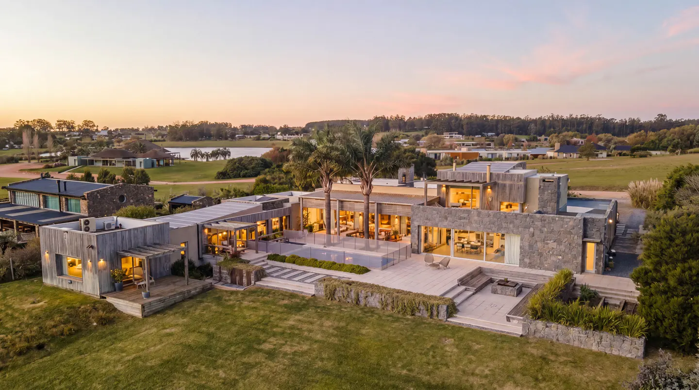Aerial view of a modern, gray stone house with palm trees and a green lawn at sunset.
