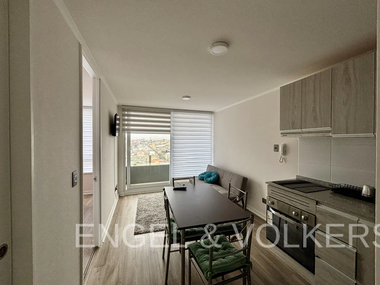 A bright apartment interior with a dining table, sofa, and kitchen area. A large window overlooks a cityscape.