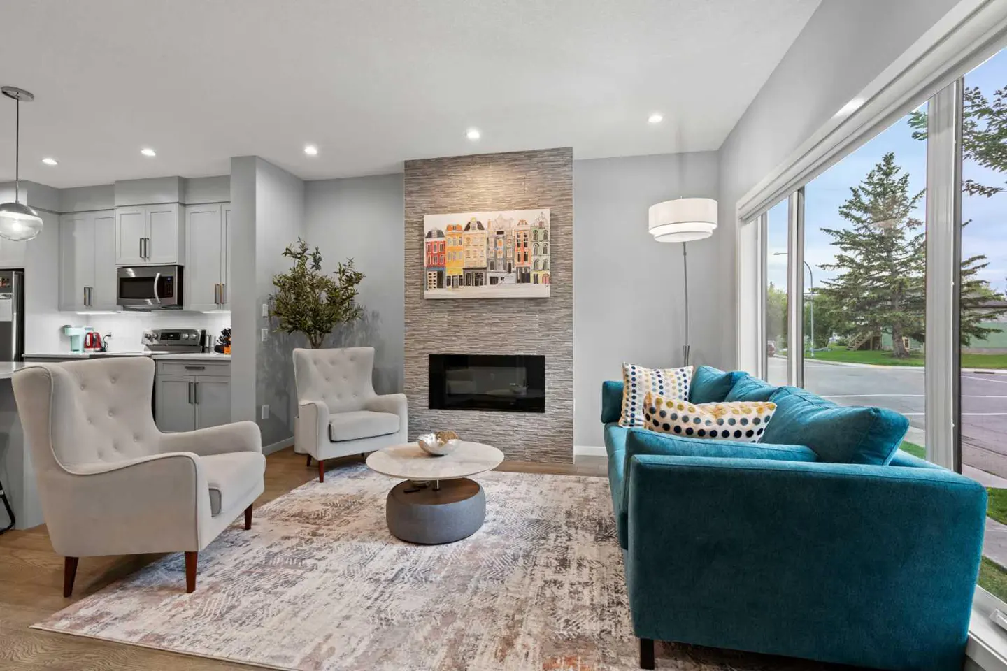 Living room with a teal sofa, two gray chairs, and a stone fireplace. A colorful painting hangs above the fireplace. Large windows offer a view of the outdoors.