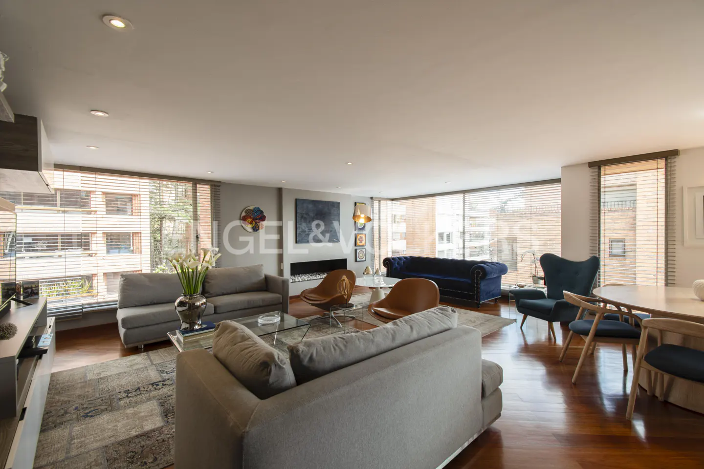 Wide shot of a living room with gray sofas, brown leather chairs, and a blue velvet couch. Hardwood floors and large windows.