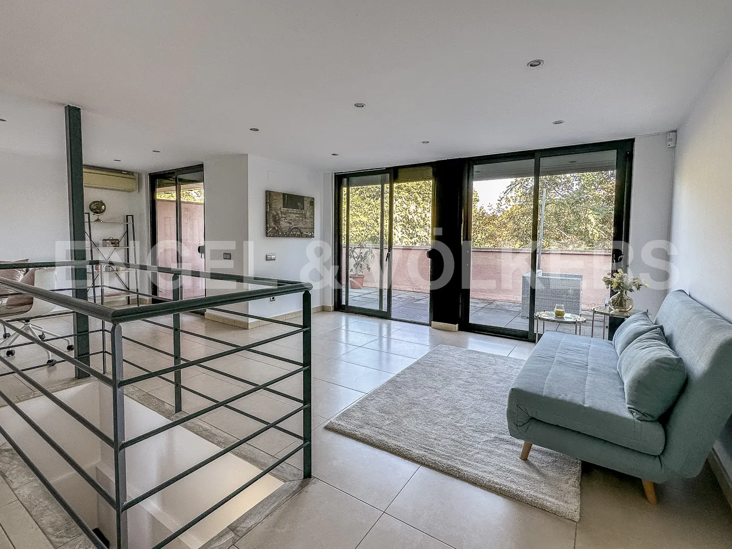 Bright, modern living room with a blue sofa, light rug, and black-framed sliding glass doors to a balcony. A staircase with dark railings is visible.