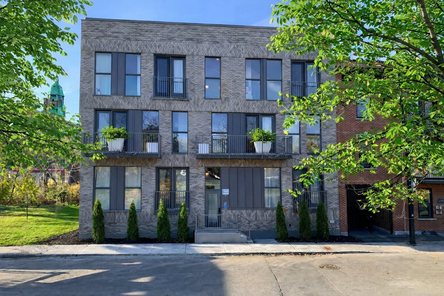 A modern, three-story apartment building with gray brick, black window frames, and small balconies. Green trees frame the building.