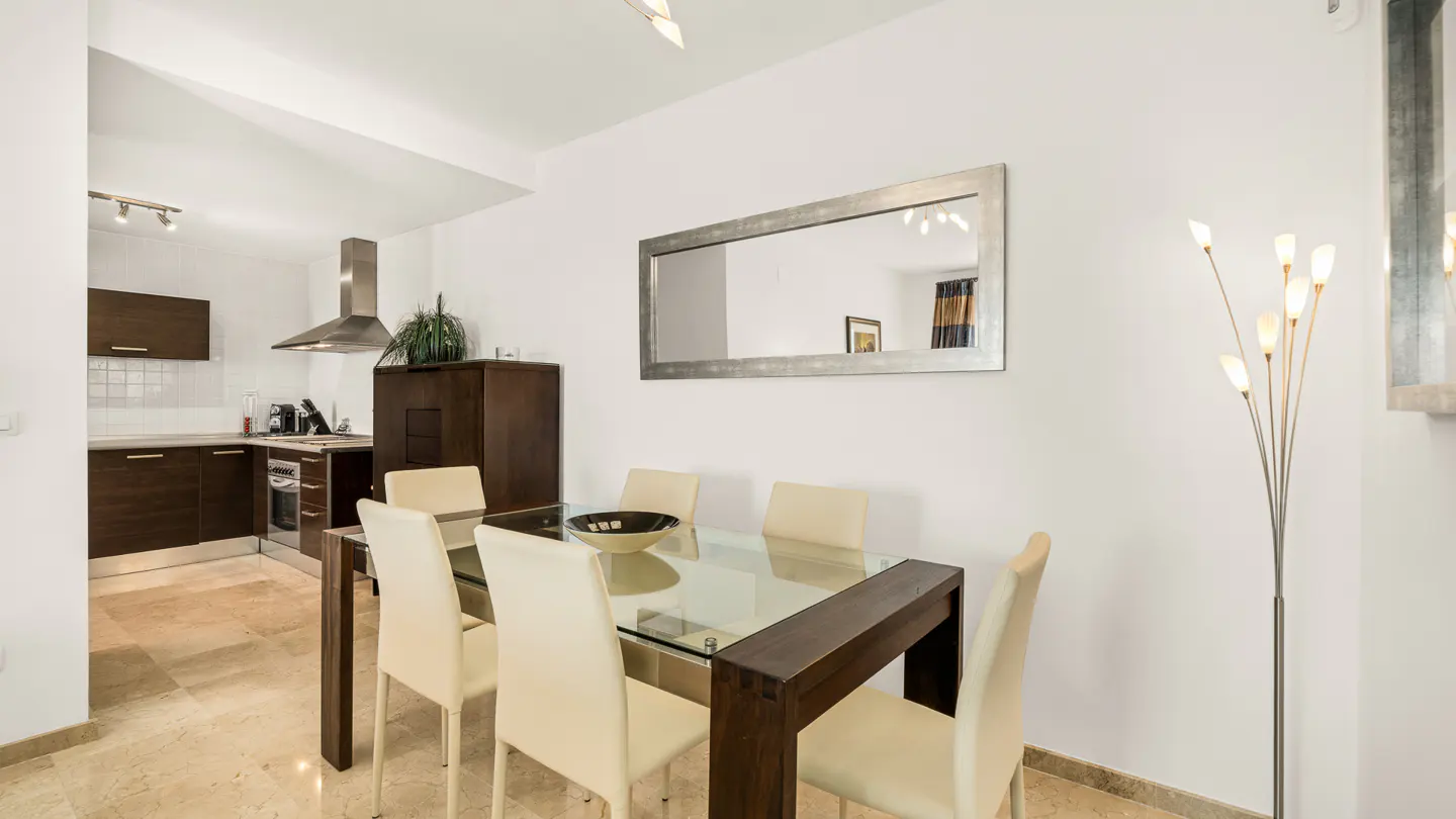Bright dining area with a glass table, cream chairs, and a dark wood frame. A mirror hangs on the white wall, reflecting the room. Kitchen in the background.