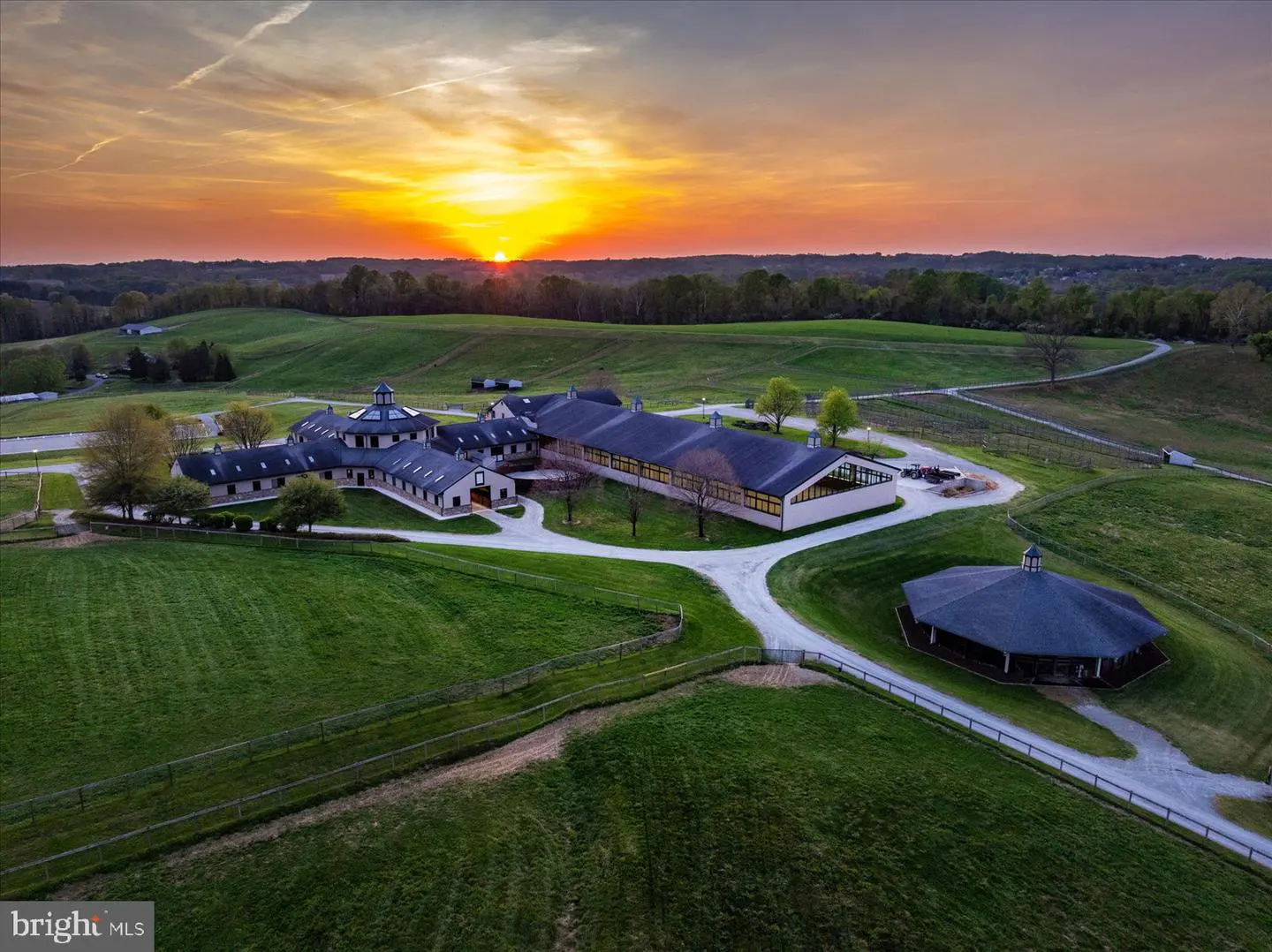 Aerial view of a horse farm at sunset, featuring green pastures, white fences, and a large barn complex with a circular building.