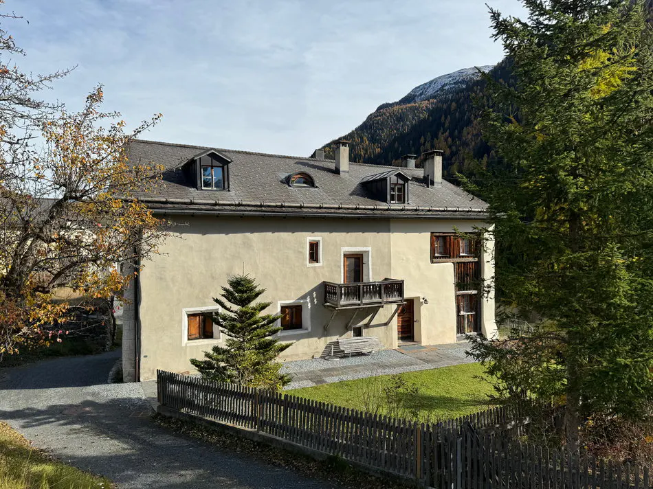 Exterior view of a beige two-story house with a gray roof, dormer windows, and a small balcony, set against a mountain backdrop.