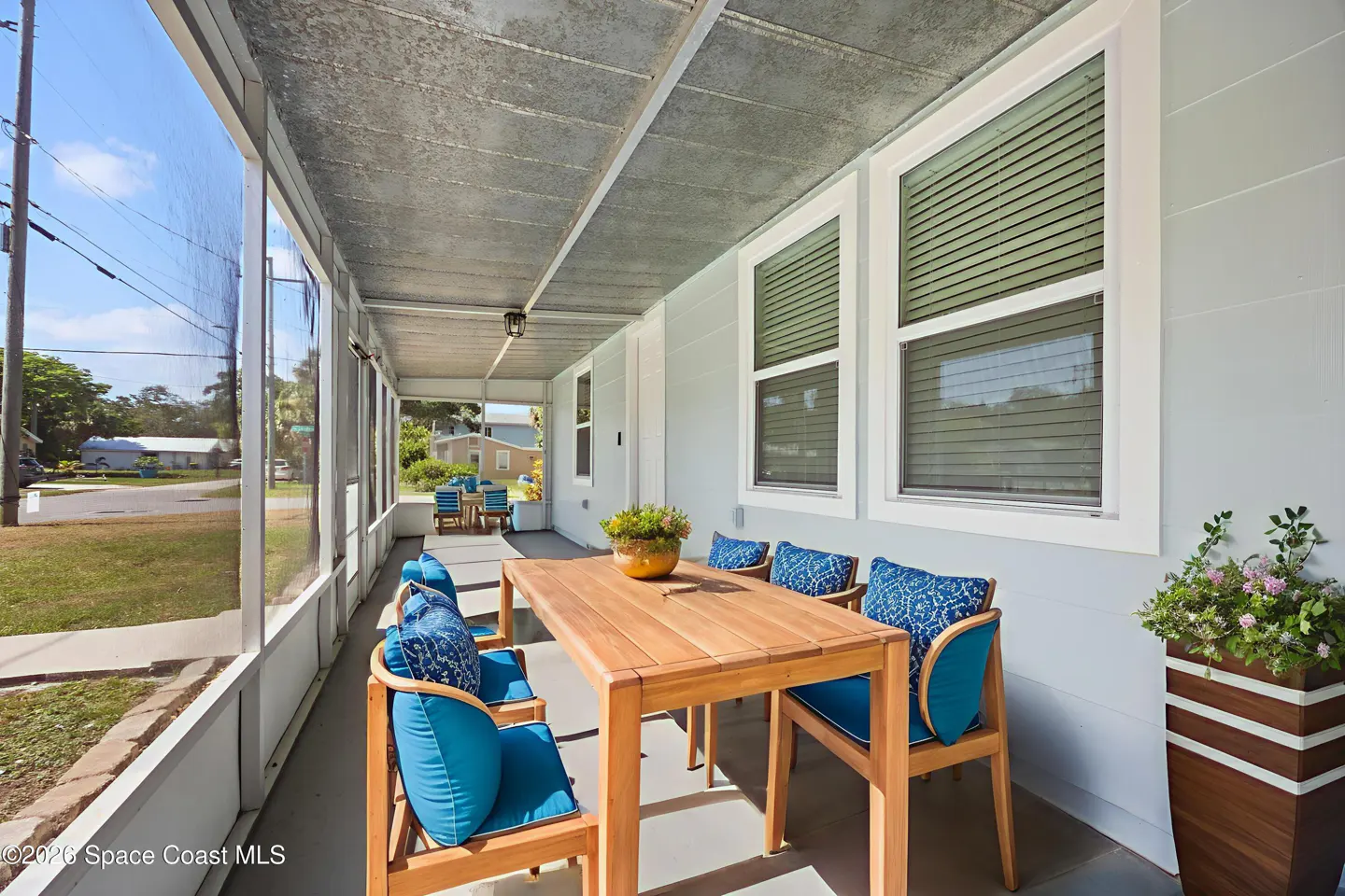 Screened porch with a wooden table and six blue cushioned chairs. Windows with blinds and a tall planter with flowers are visible.