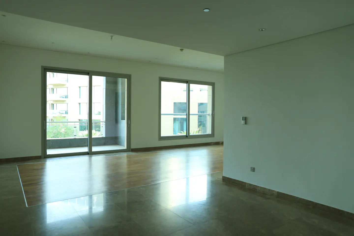 Empty room with light walls, marble floors, and two large windows with views of a building and balcony.