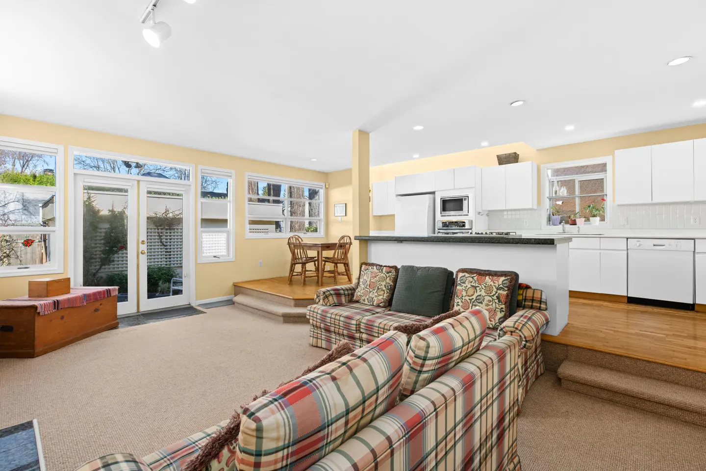 Open-concept living room and kitchen with plaid sofas, white cabinets, and light yellow walls. Windows and doors lead to the outside.