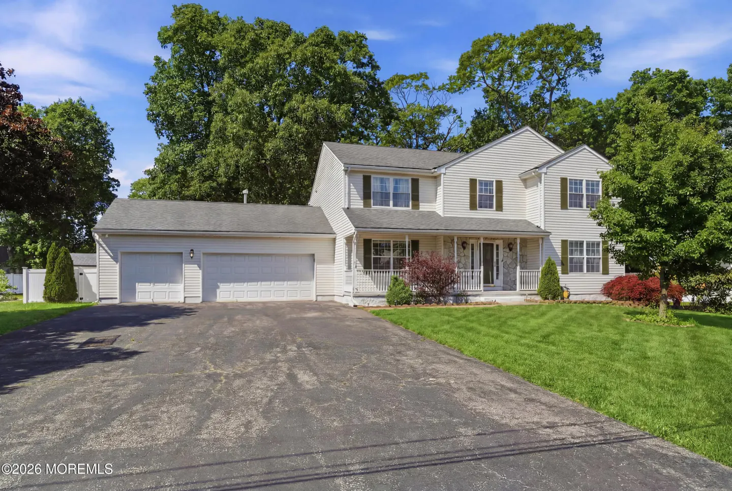 Two-story beige colonial home with a three-car garage, front porch, and large green lawn under a blue sky.
