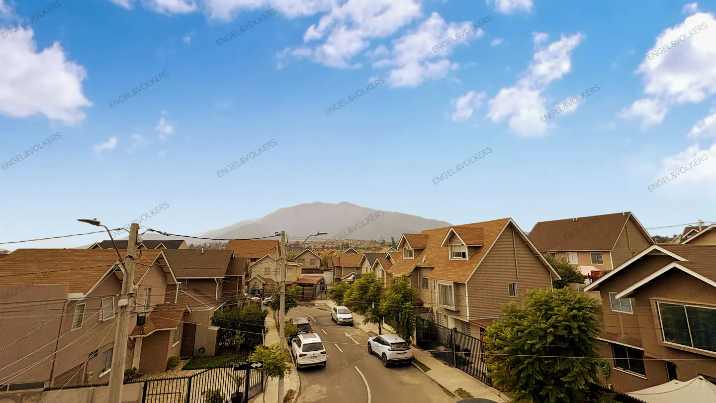 Residential street view with brown houses, cars, and a mountain backdrop under a blue sky with white clouds.