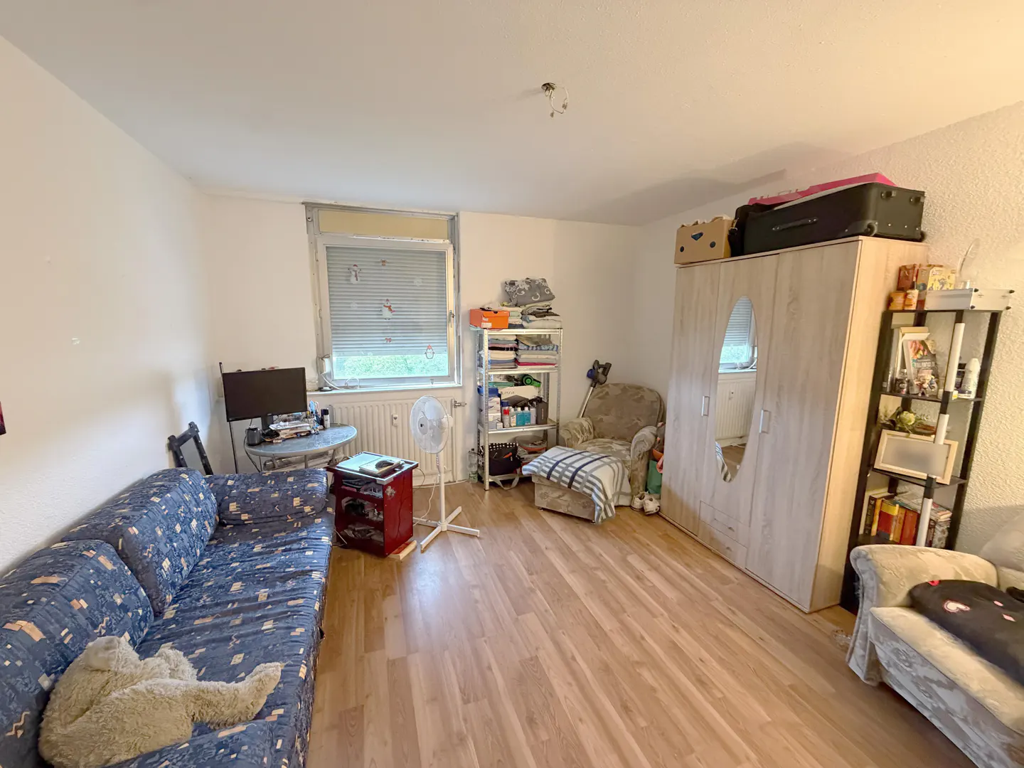 A wide shot of a furnished room with a blue patterned sofa, wooden floors, and a large wardrobe. A window with blinds is on the left.