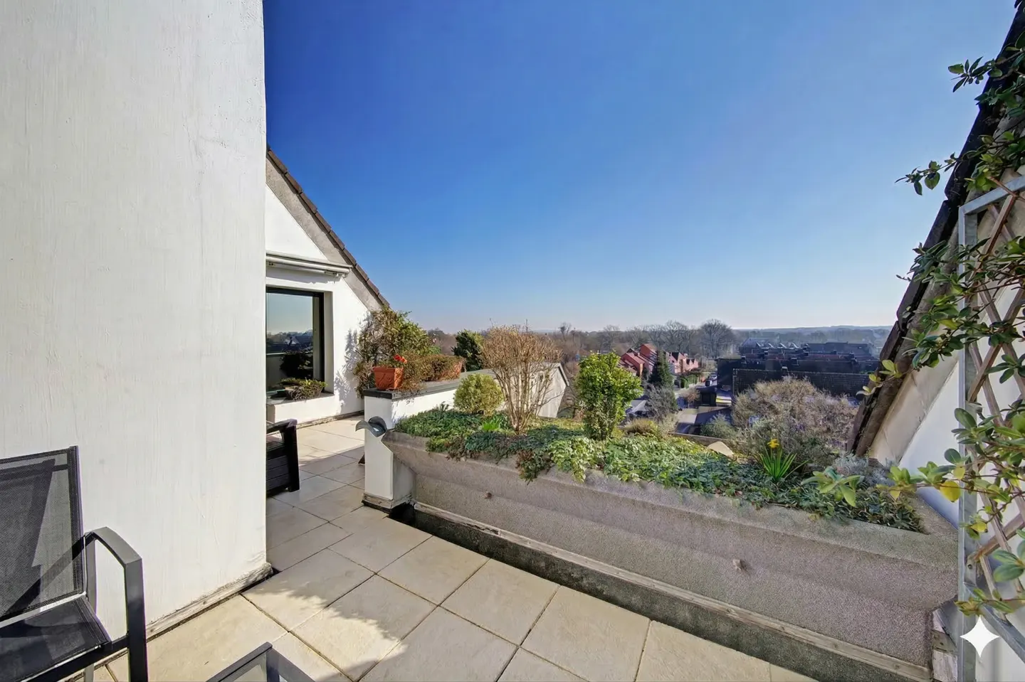 A rooftop patio with a concrete planter filled with greenery overlooks a neighborhood under a clear blue sky.
