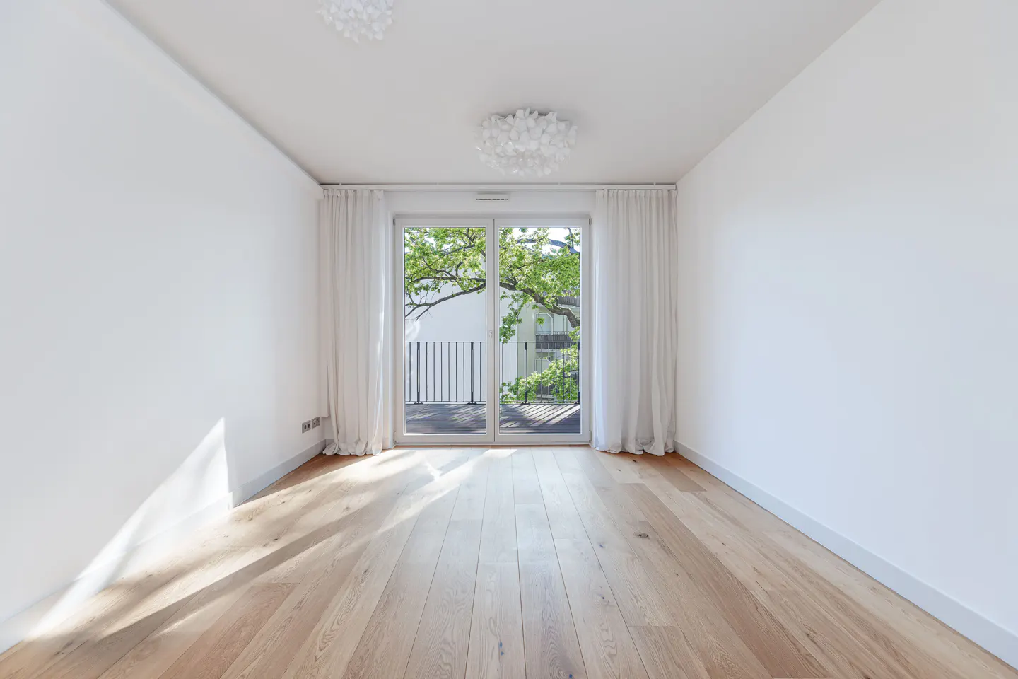 Bright, empty room with light wood floors, white walls, and glass doors to a balcony with green trees visible.