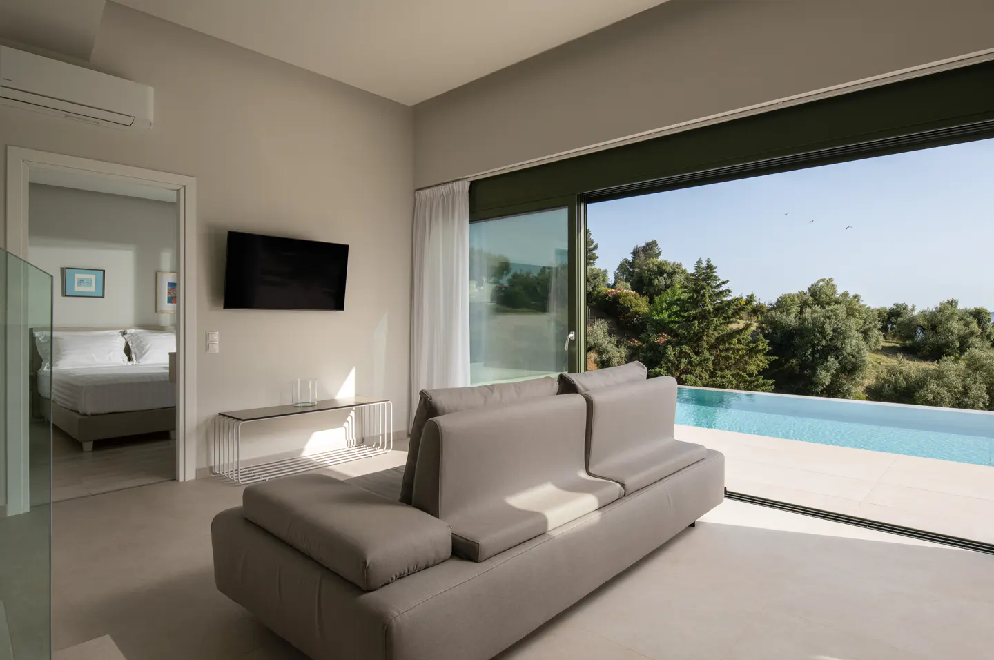 Living room with gray sofa, TV, and glass table. Sliding glass doors open to a pool and green landscape. Bedroom visible through doorway.