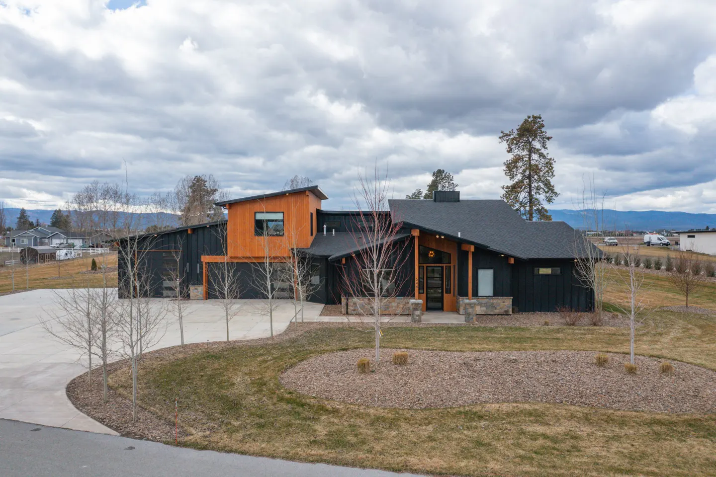 Modern black house with wood accents, a dark roof, and a large driveway. The lawn is green with brown mulch. Cloudy sky.