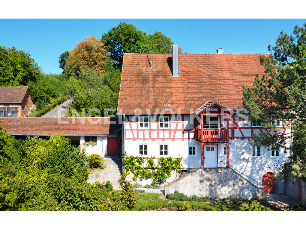Traditional half-timbered house with a red tile roof, white walls, and a small balcony, surrounded by lush green trees. [AI-generated text]
