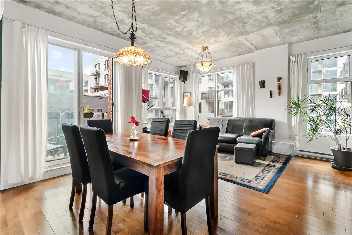 Bright living room with wood floors, a dining table with black chairs, and a gray sofa. Large windows with white curtains offer city views.