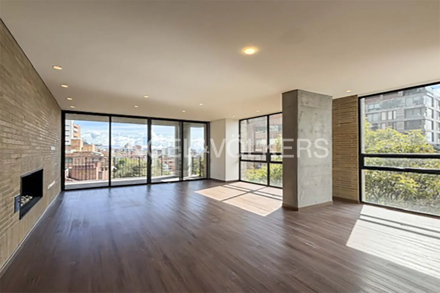 Spacious, bright living room with hardwood floors, brick accent wall, and large windows offering city views.