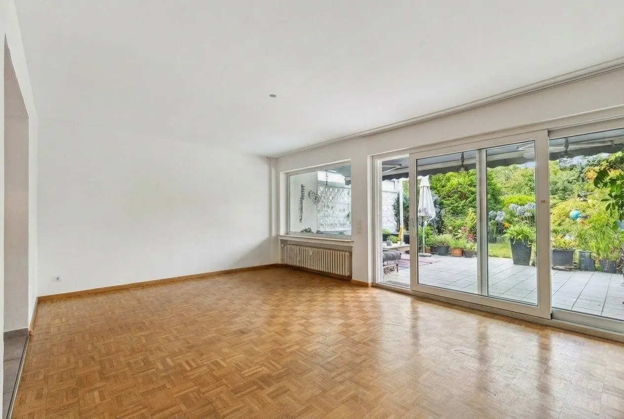 Bright, empty room with parquet floors, white walls, and sliding glass doors to a patio with plants.