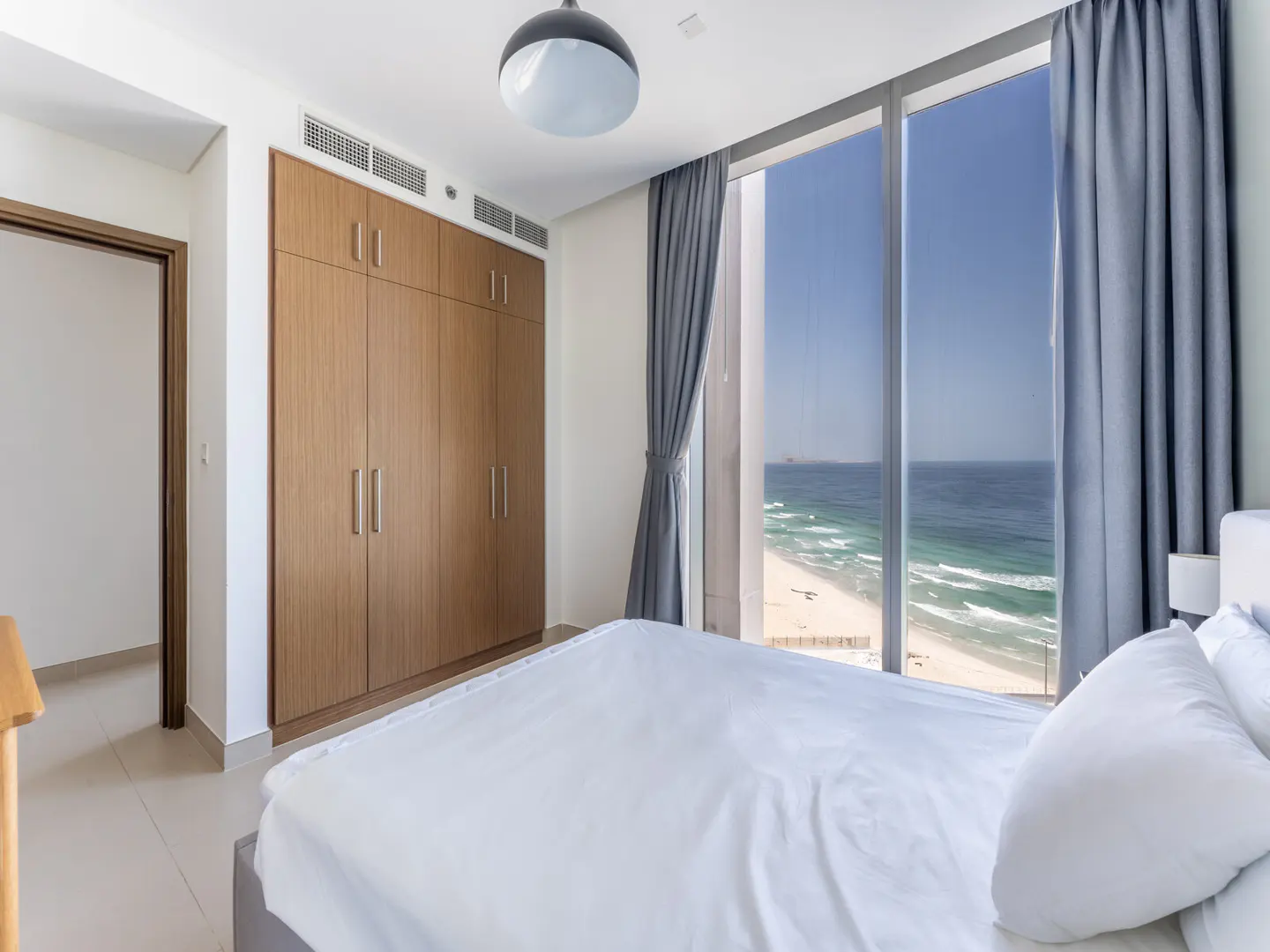 Bedroom with a white bed, wood closet, and a large window with ocean view and blue curtains.