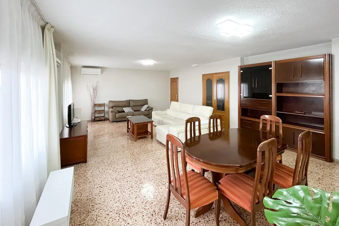 Bright living room with a brown dining table, chairs, a white sofa, and a brown sofa. A large wooden cabinet stands against the wall.