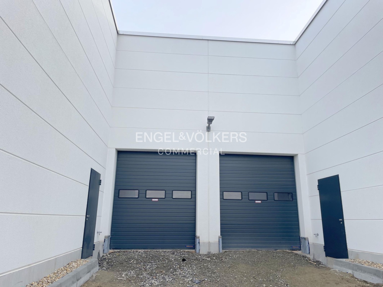 Exterior view of a modern industrial building with two dark grey roll-up garage doors and two side entry doors. [AI-generated text]