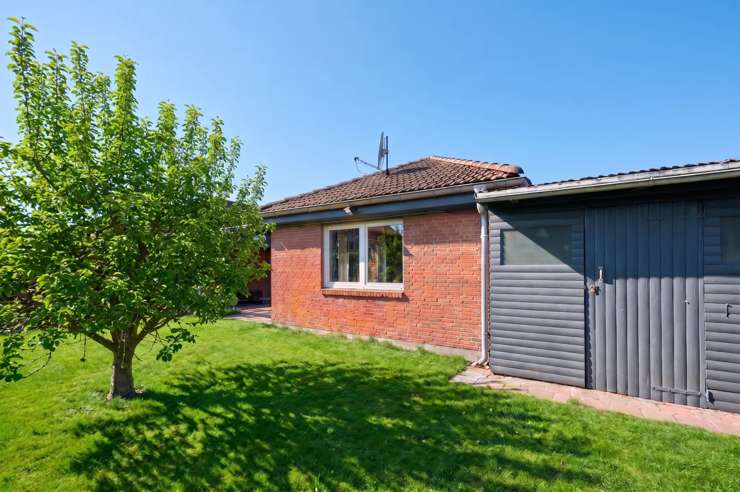 Red brick house exterior with a green lawn, a leafy tree, and a dark grey wooden shed under a clear blue sky.