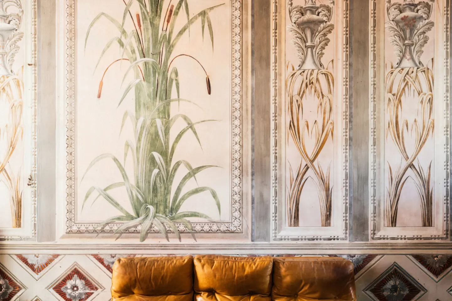 Ornate wall with painted cattails and floral designs above a brown leather sofa and patterned tile base.