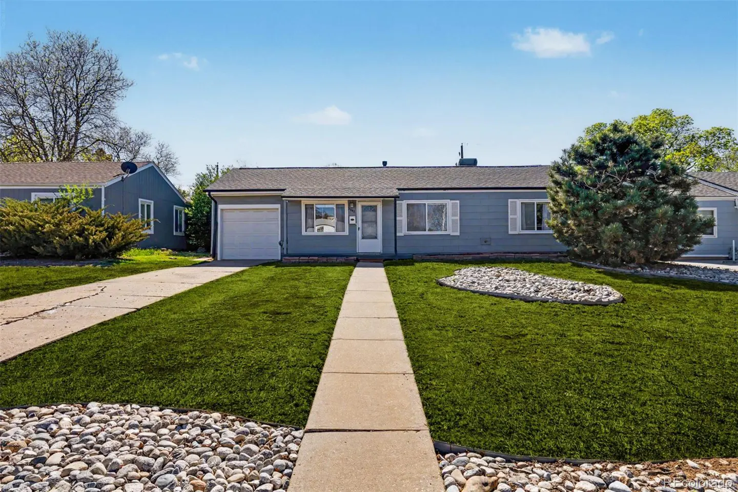 A single-story, light blue house with white trim and a green lawn under a blue sky. A concrete walkway leads to the front door.