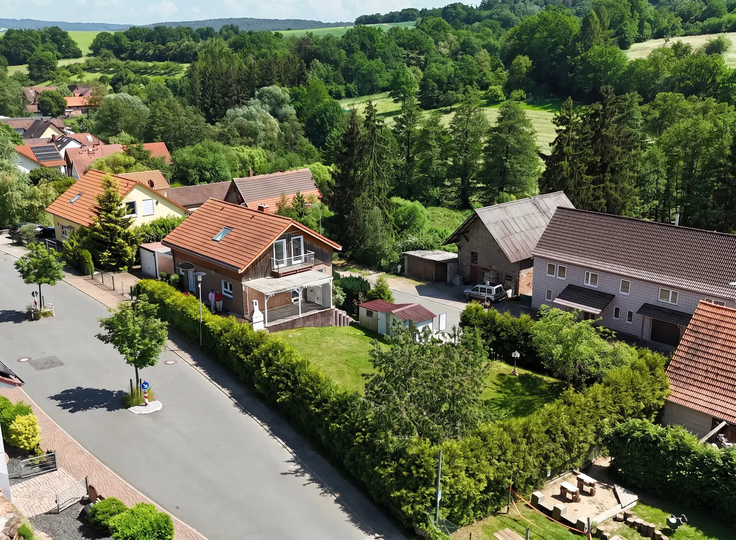 Aerial view of a quiet street with houses, green trees, and a forest in the background. The houses have red tile roofs.