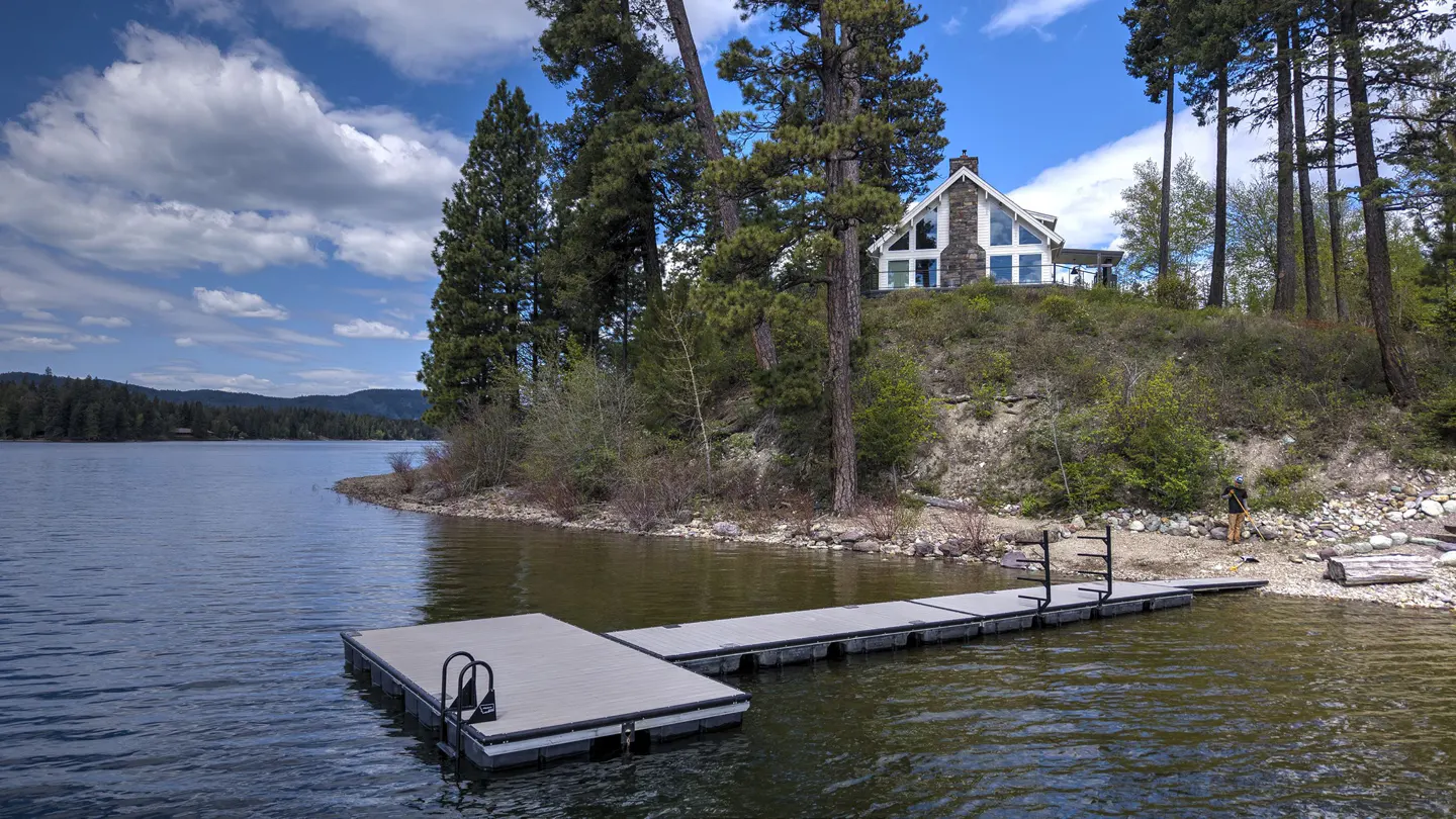 Lakefront home with a dock. A modern house sits on a hill with tall trees. Blue sky with clouds. Person on the beach.
