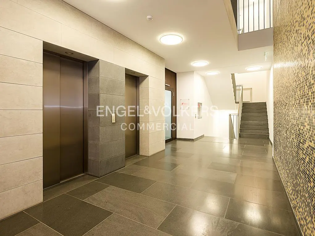 Modern lobby with two elevators, gray tiled floor, and a staircase in the background. The wall on the right is decorated with mosaic tiles.