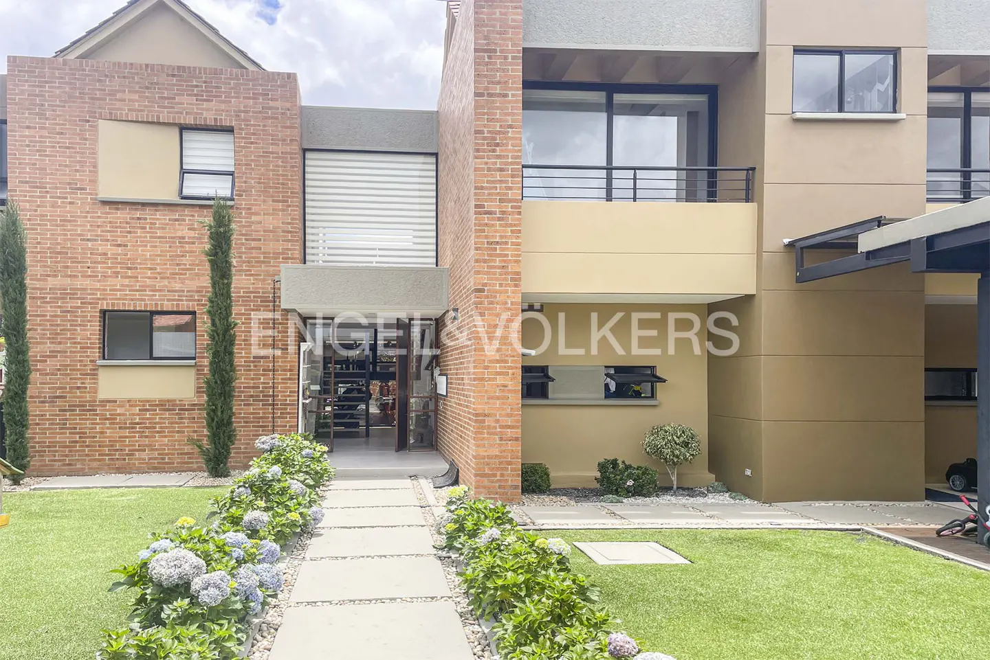 Exterior view of a modern brick and tan townhouse with a stone walkway, green lawn, and blue hydrangeas. Engel & Volkers logo on the building.